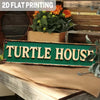A vintage metal sign with the text TURTLE HOUSE is displayed on a wooden surface next to potted plants.