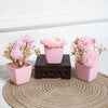A set of three pink artificial rose potted plants displayed on a wooden block and a woven mat.