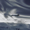 A military jet aircraft flying through swirling clouds above a snowy mountain landscape.