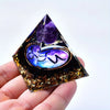 A hand holds a dark pyramid crystal with purple and gold inclusions, shaped like a triangle.