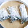 A collection of white cotton candle wicks with metal tips arranged on a light wooden surface.