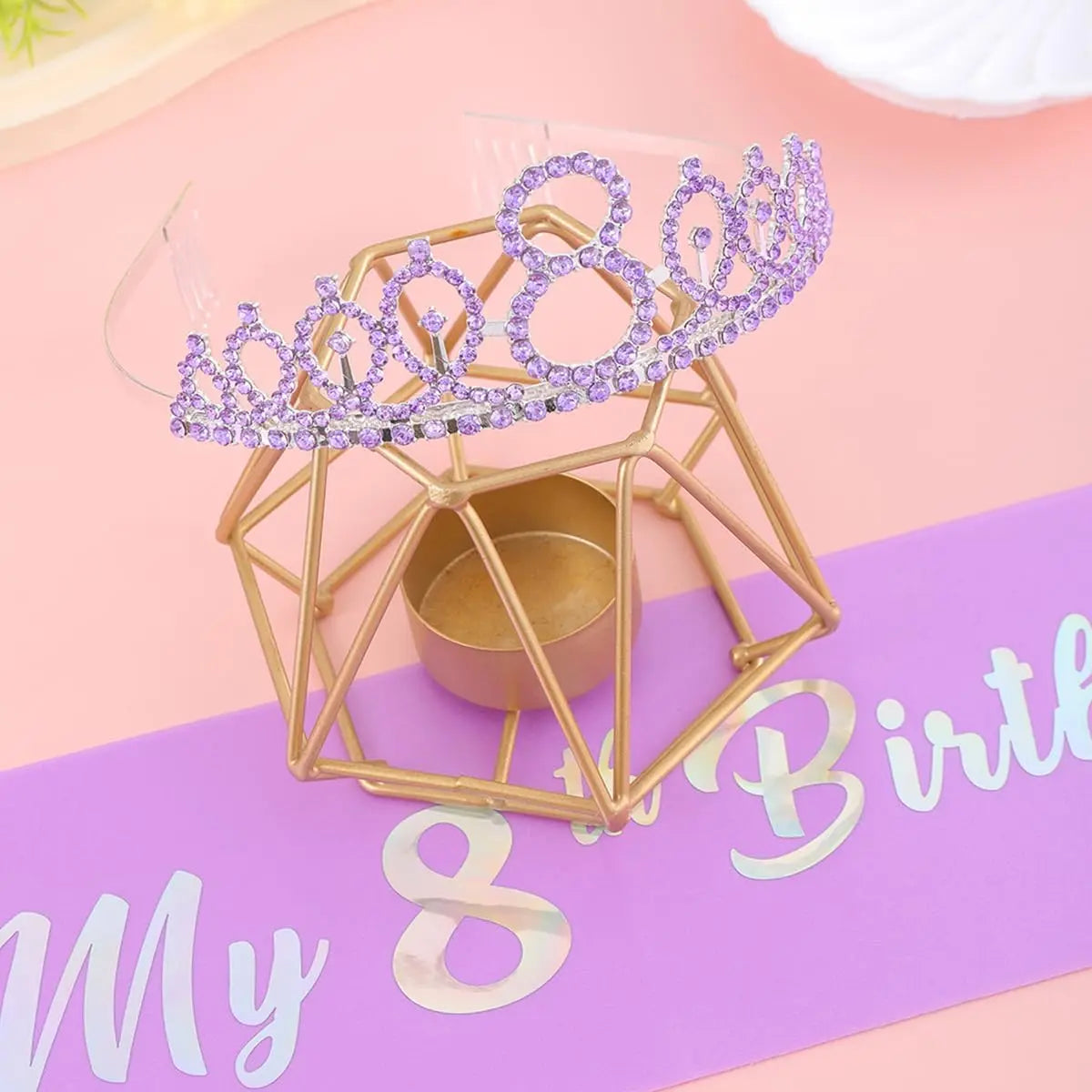 A gold geometric crown structure adorned with purple sparkling gems and a small gold container, set against a pink background