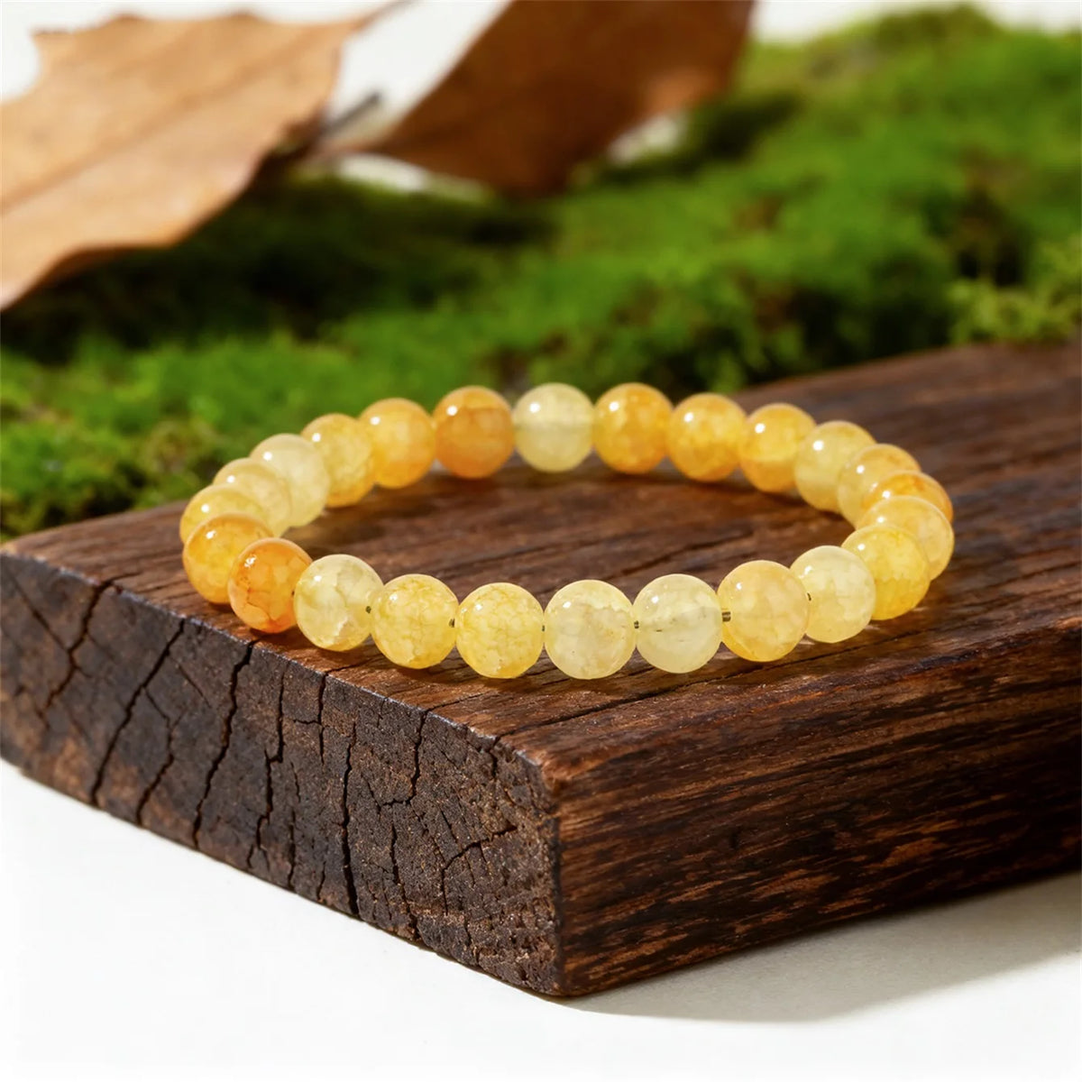 A bracelet of small, round, yellow-orange citrine crystals is displayed on a dark wooden surface with green moss in the backg