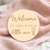 A round wooden sign with gold lettering that reads Welcome to the world little one placed on a pink knitted fabric.