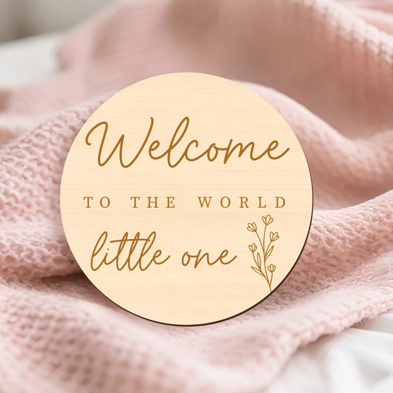 A round wooden sign with gold lettering that reads Welcome to the world little one placed on a pink knitted fabric.