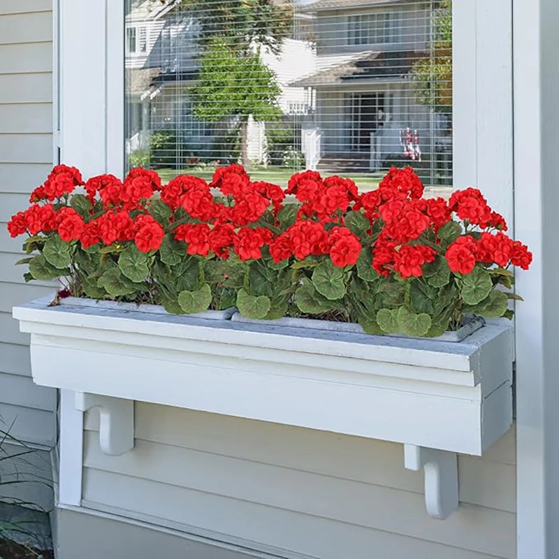 A planter filled with bright red silk faux geranium flowers sits on a white wooden ledge.