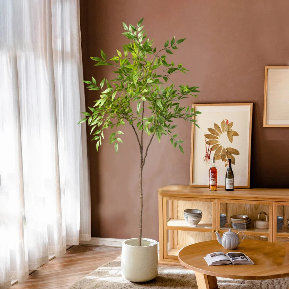 A tall artificial Nandina plant with green leaves sits in a white pot on a wooden table next to a framed picture.