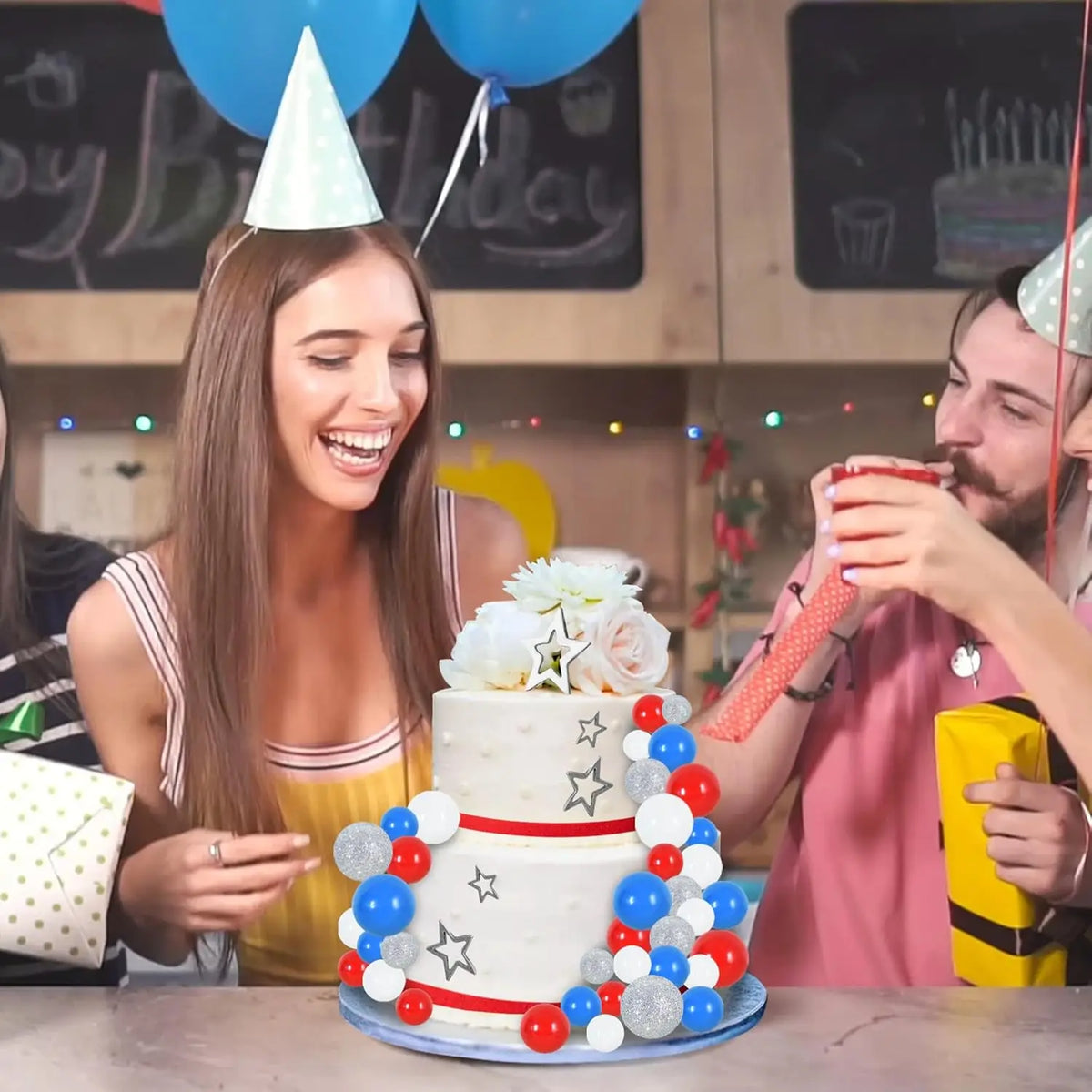 A woman smiles while decorating a multi-tiered white cake with colorful foam ball toppers in blue, red, and silver.