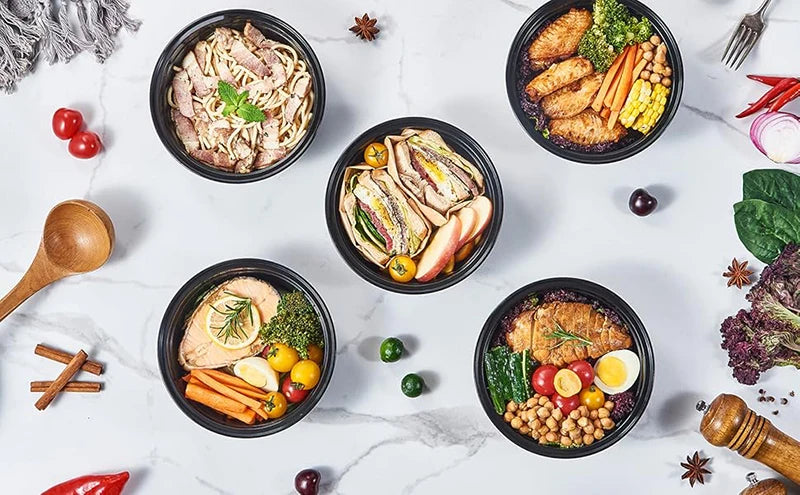 A variety of healthy meal prep foods including sliced meat, vegetables, and grains are displayed in black round containers on