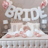 A bridal shower decoration featuring white letter foil balloons spelling BRIDE above a bed with pink and white accents.