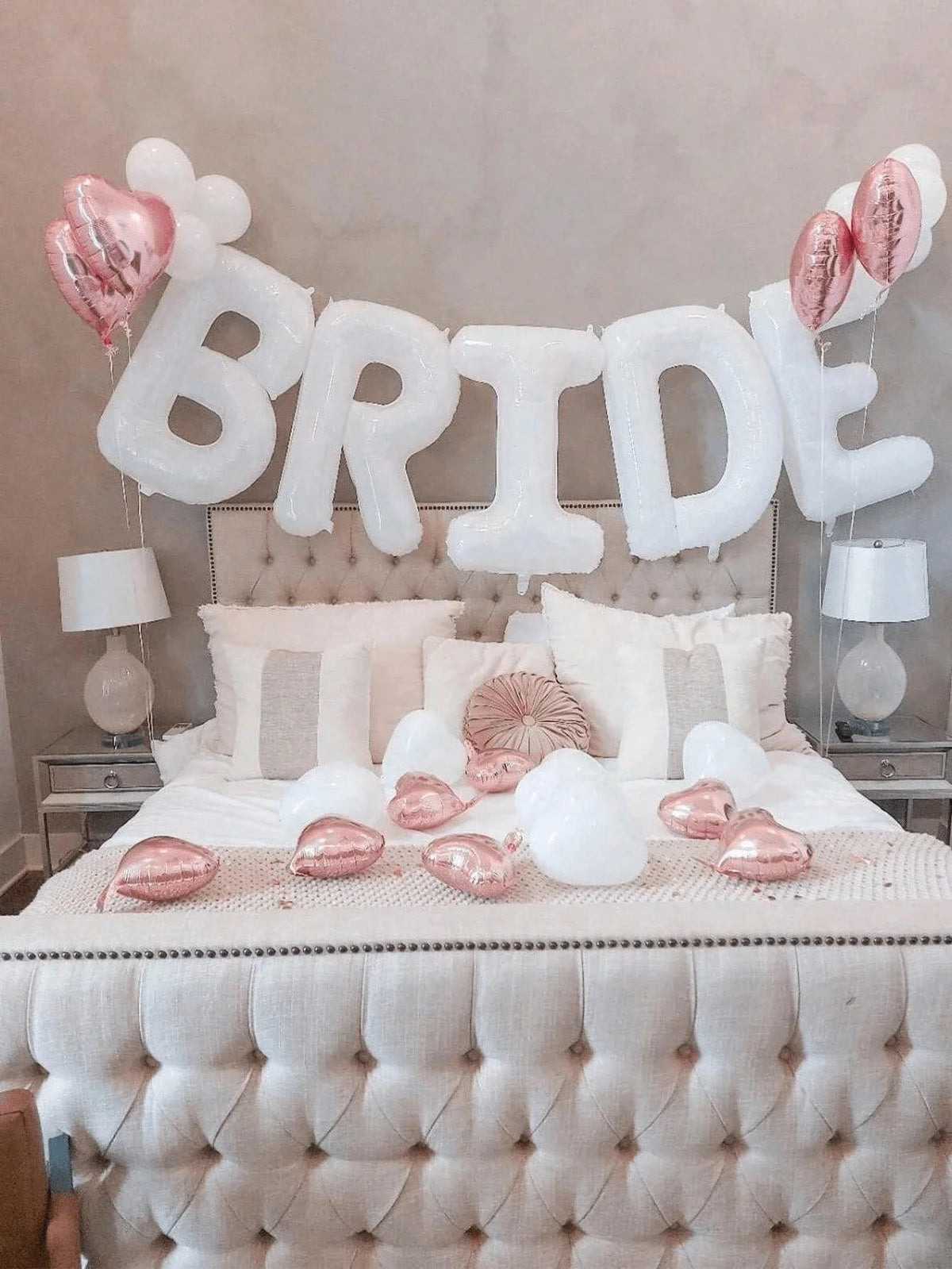 A bridal shower decoration featuring white letter foil balloons spelling BRIDE above a bed with pink and white accents.