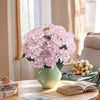 A bouquet of pink artificial hydrangea flowers in a green vase on a wooden table next to an open book.