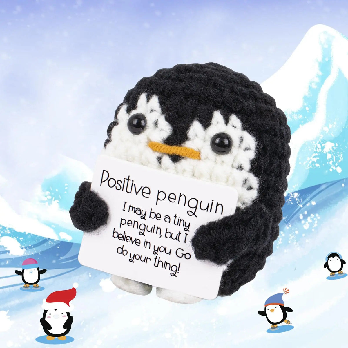A black and white crocheted penguin plush holding a card with an encouraging message in a snowy outdoor setting.