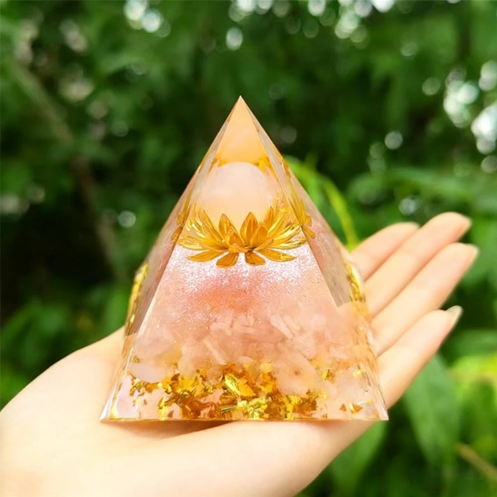 Handmade resin orgonite pyramid with natural pink quartz and gold inclusions held against a green leafy background