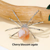 A handcrafted crystal spider figurine with silver legs and a cherry blossom agate body resting on a light background.