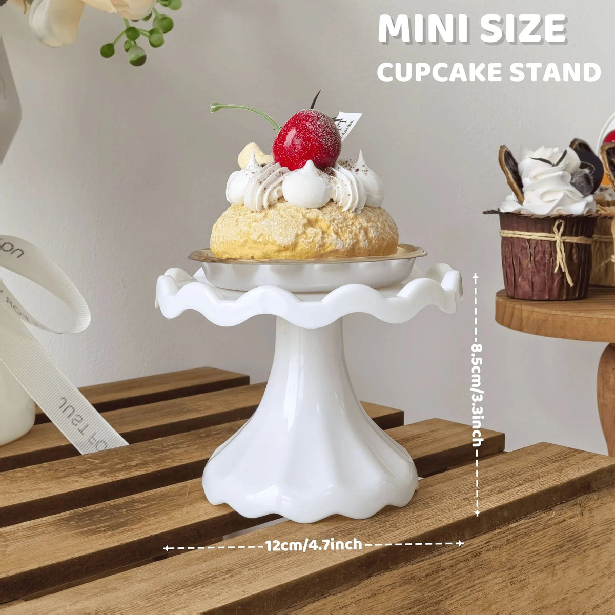 A white mini cupcake stand displaying a single cake topped with a red berry and whipped cream on a wooden surface.