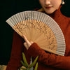 A woman in a rust-colored sweater holds an elegant bamboo folding fan with delicate leaf designs.