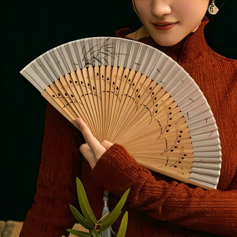 A woman in a rust-colored sweater holds an elegant bamboo folding fan with delicate leaf designs.