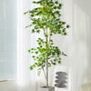 A tall artificial maple tree with silk green leaves stands in a gray planter next to a window.