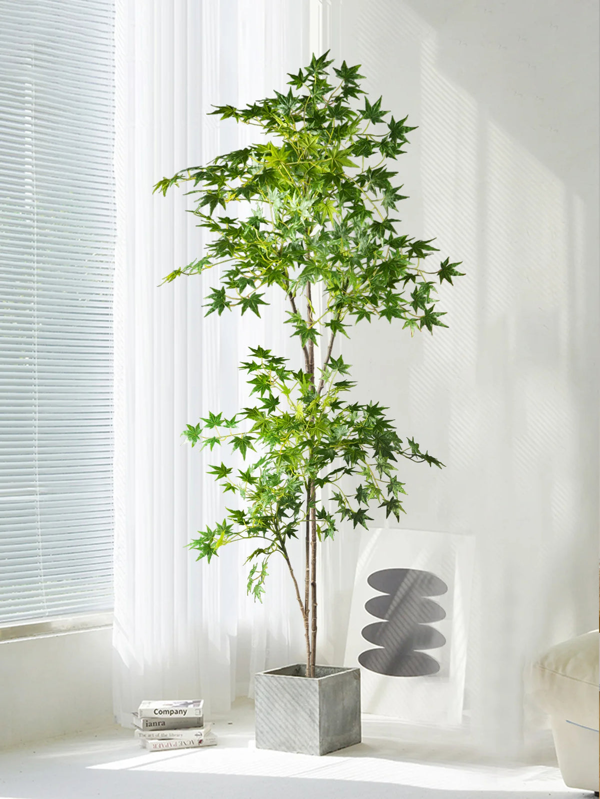 A tall artificial maple tree with silk green leaves stands in a gray planter next to a window.
