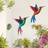 Vibrant metal hummingbird wall art featuring iridescent colors displayed against a white wall with various potted plants.