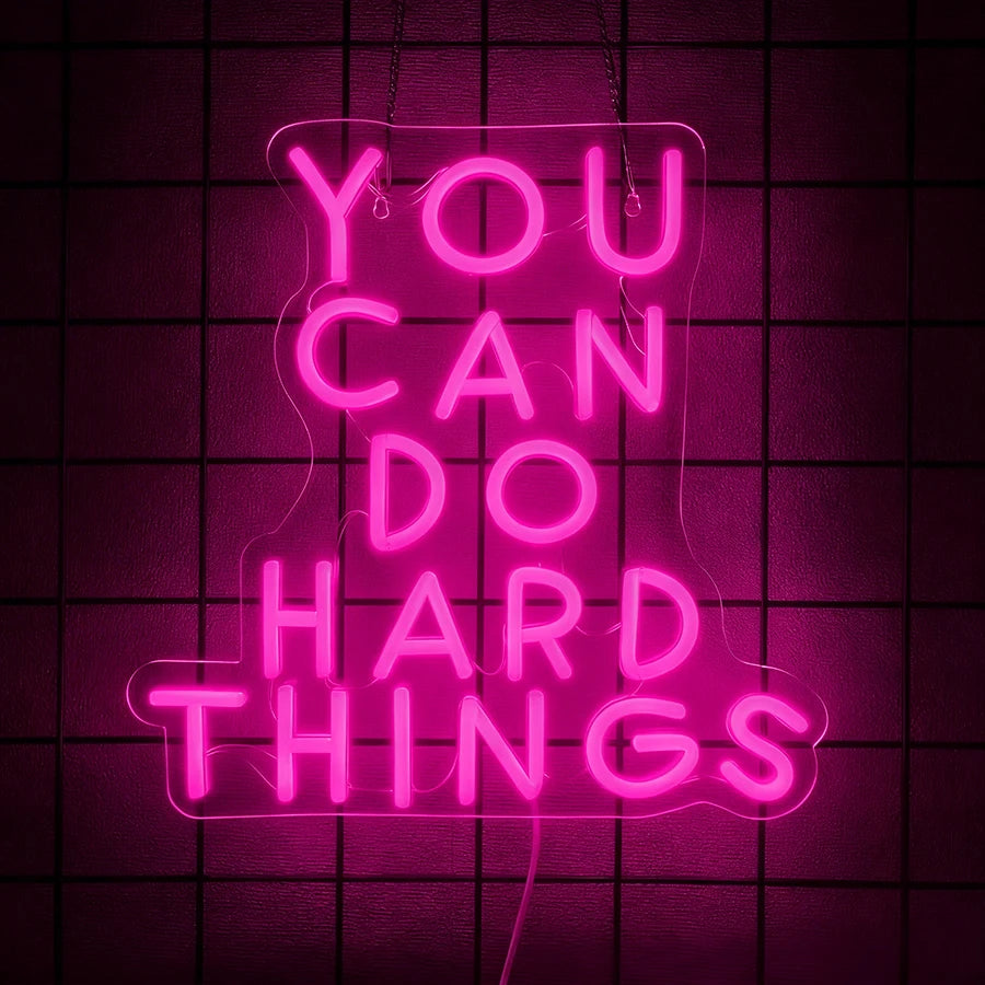 Pink neon sign shaped like a bell with the text YOU CAN DO HARD THINGS displayed against a dark grid background.