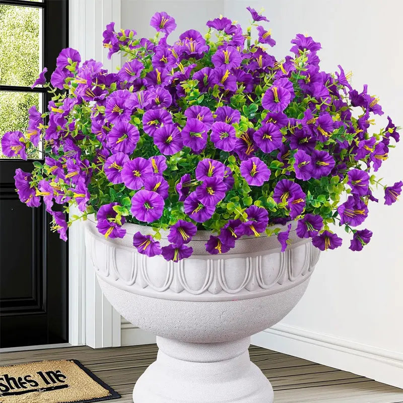 A vibrant bouquet of purple artificial silk cloth flowers displayed in a white urn on a wooden floor.
