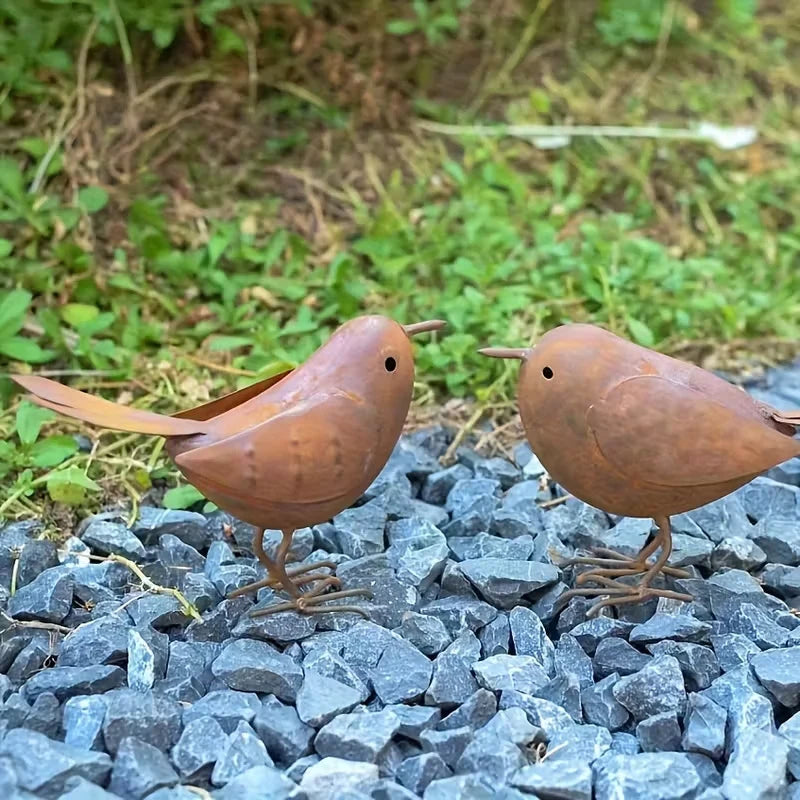 Two rustic metal bird sculptures standing on dark gray gravel with green foliage in the background.