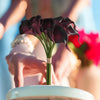 A bouquet of six dark purple artificial calla lilies held by a person against a bright blue background.