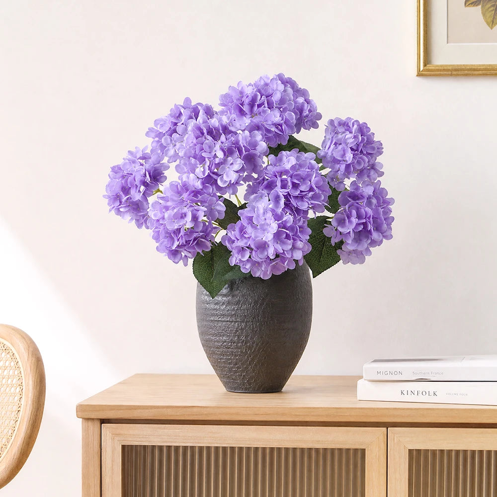 A bouquet of purple artificial hydrangea silk flowers in a dark gray vase on a wooden table.