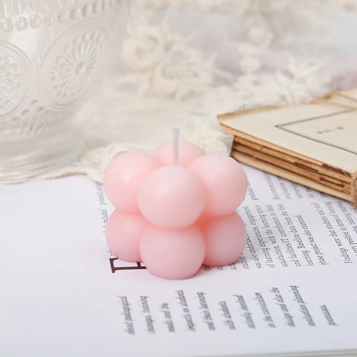 A set of two pink cube soy wax candles resting on a white notebook next to lace fabric.