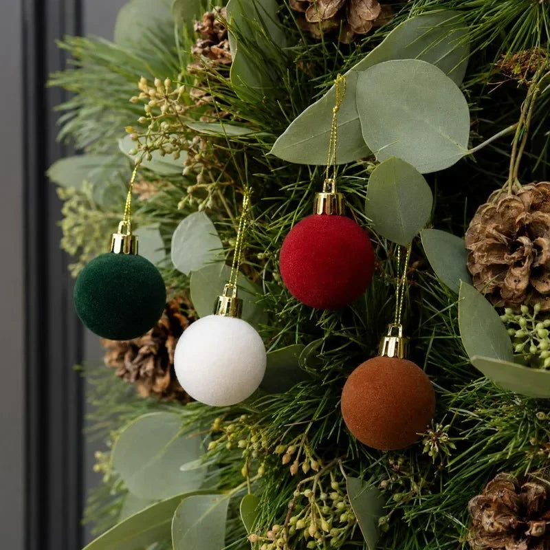 A close-up of five velvet flocked Christmas tree ornaments in shades of green, red, white, and copper hanging on a fir branch