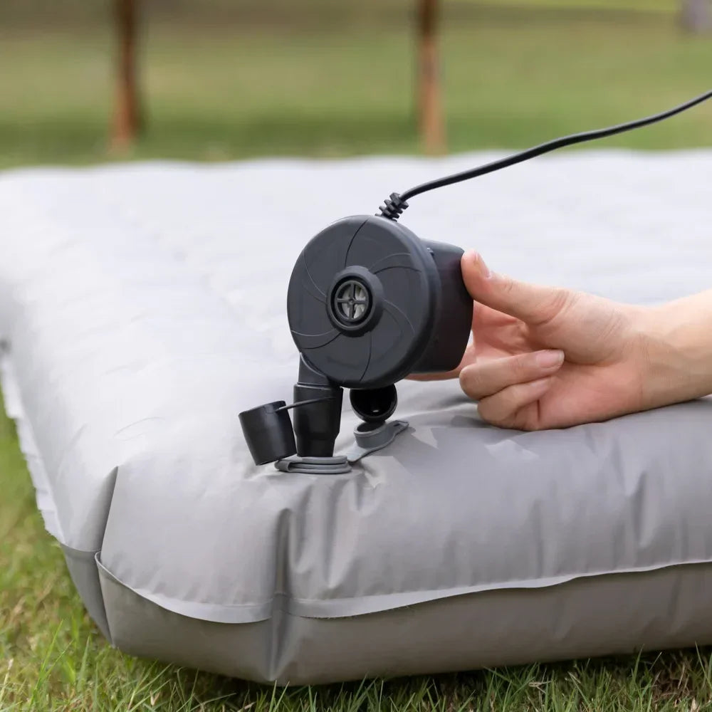 Eine Hand verbindet eine portable elektrische Luftpumpe mit einem aufblasbaren Gegenstand auf einer grauen Matte im Freien.
