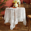 Elegant white floral lace tablecloth covering a wooden table set with yellow flowers and glass