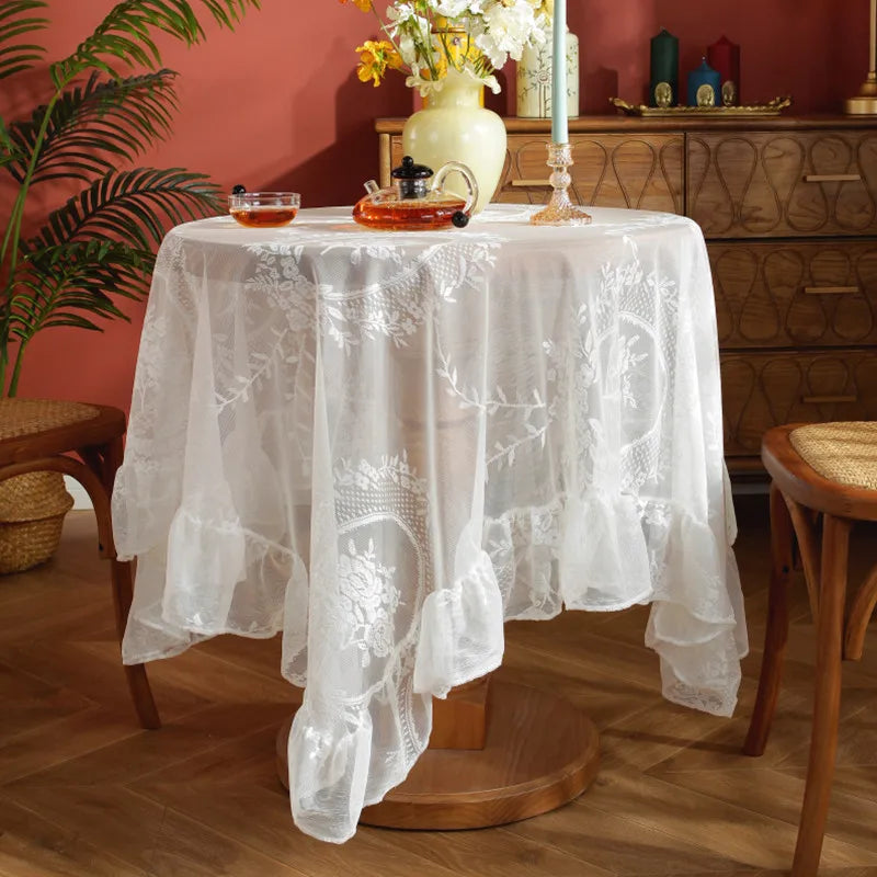 Elegant white floral lace tablecloth covering a wooden table set with yellow flowers and glass