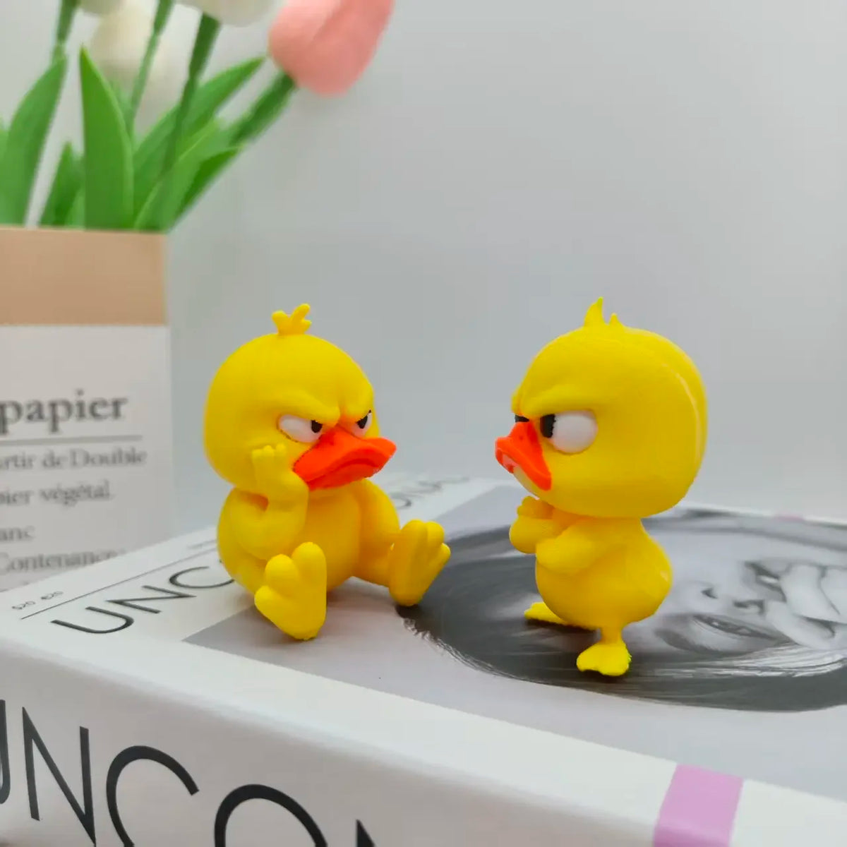 A yellow 3D printed angry duck figurine sitting on a white box with a floral background.