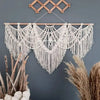 Handwoven boho macrame wall hanging tapestry with fringe in off-white color against a gray wall.