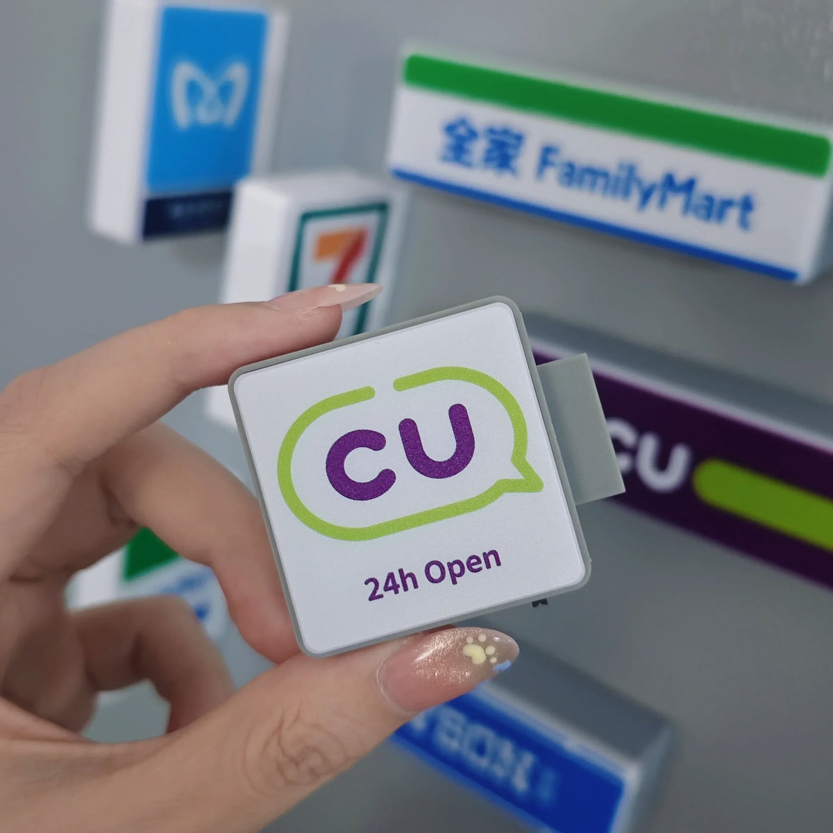 A hand holds a white fridge magnet sign with green and purple text indicating 24h open for FamilyMart.