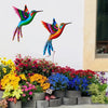 Vibrant metal hummingbird wall art featuring iridescent colors displayed against a white wall with colorful garden plants bel