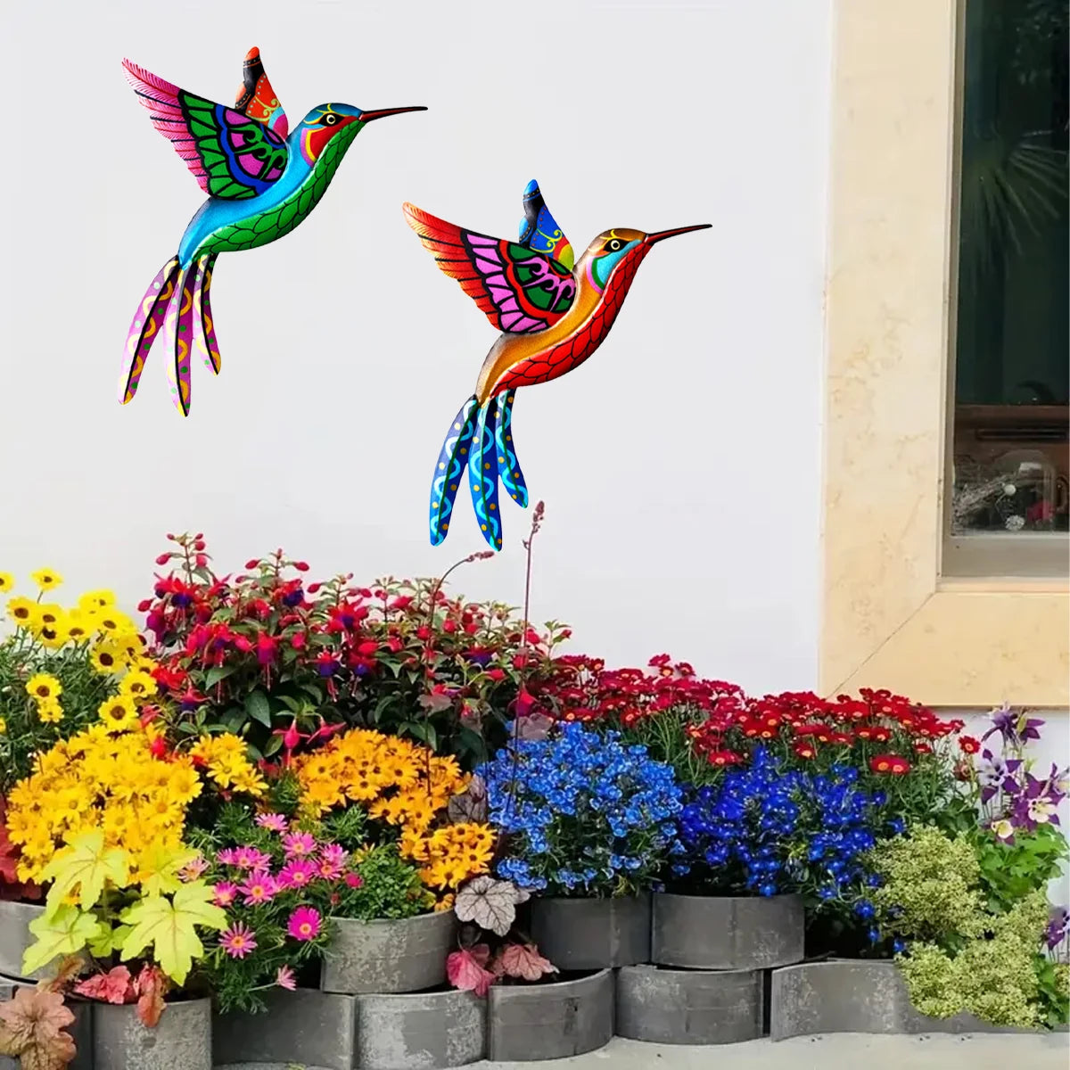 Vibrant metal hummingbird wall art featuring iridescent colors displayed against a white wall with colorful garden plants bel