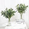 A glass vase filled with artificial olive leaf branches on a white table.