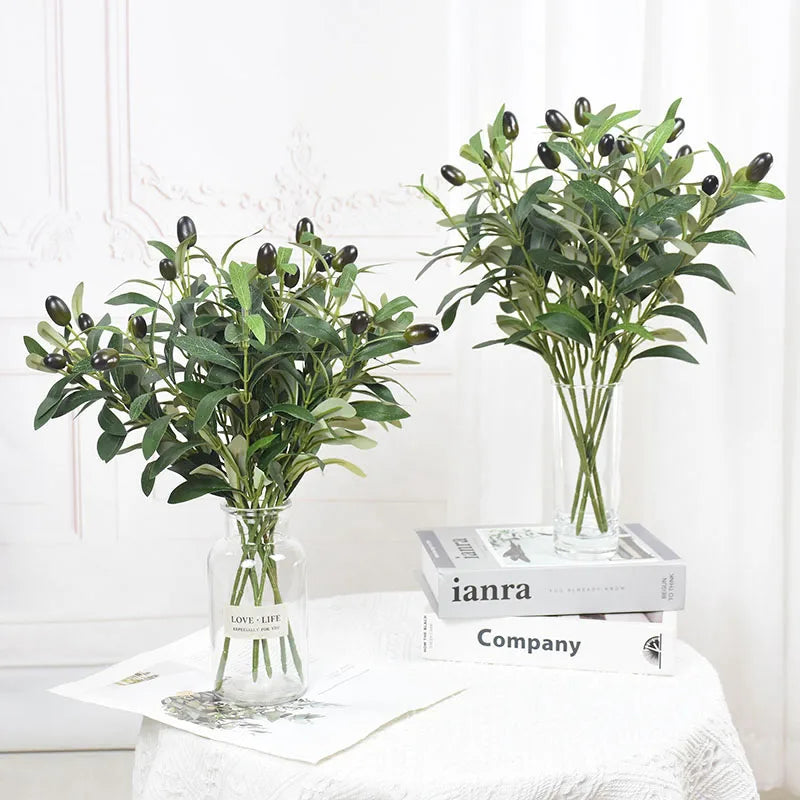 A glass vase filled with artificial olive leaf branches on a white table.