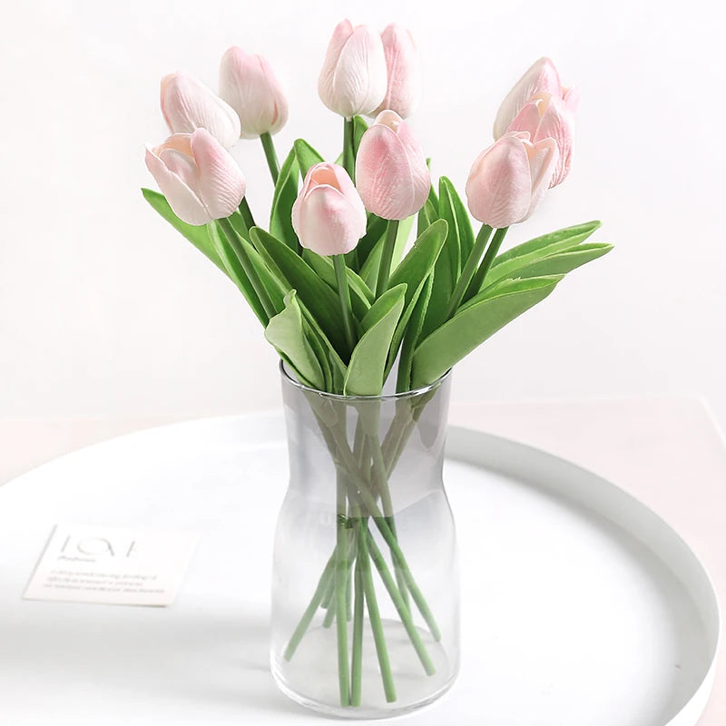 A bouquet of pink and white artificial tulip flowers in a clear glass vase on a white plate.