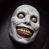 A realistic smiling demon latex mask with pale gray skin and menacing teeth against a dark background.