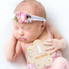 A newborn baby sleeping peacefully wearing a pink floral headband and holding a wooden first month milestone card.