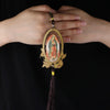 A hand holds a wooden Our Lady of Guadalupe pendant charm with a gold-colored loop and a dark tassel.