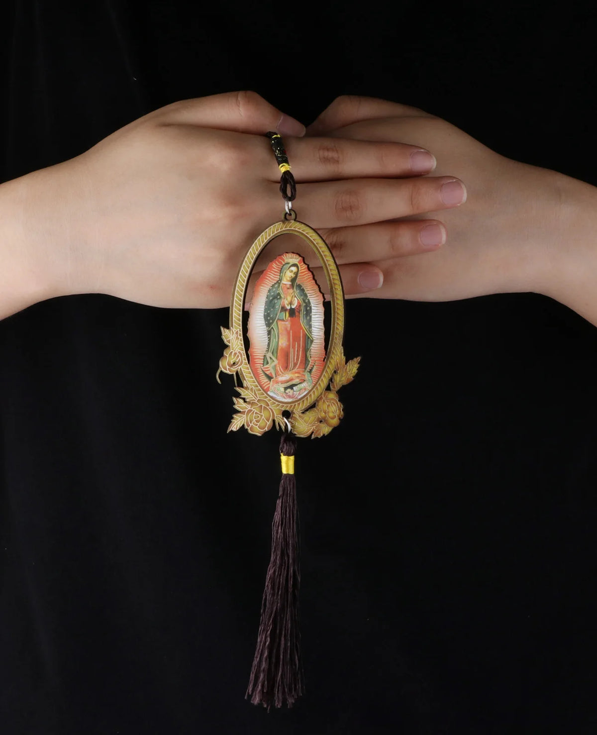 A hand holds a wooden Our Lady of Guadalupe pendant charm with a gold-colored loop and a dark tassel.