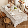 An elegant white floral lace tablecloth set for a dining table with silverware and fruit.