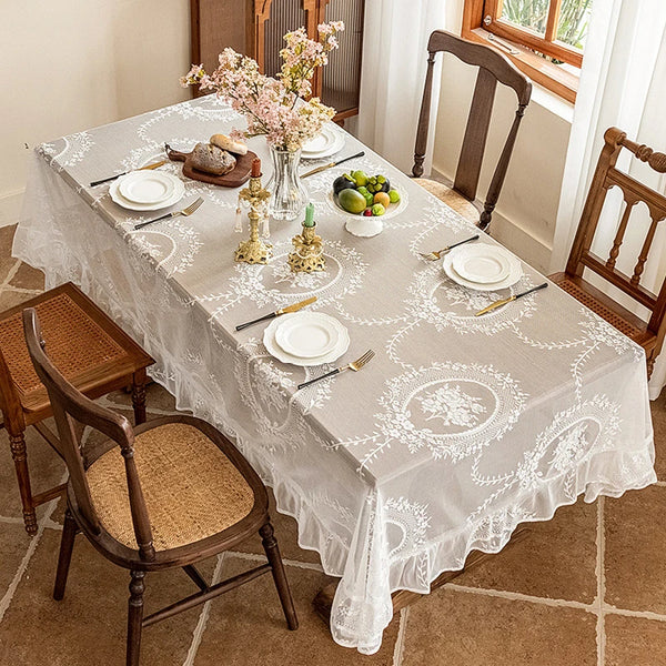 An elegant white floral lace tablecloth set for a dining table with silverware and fruit.