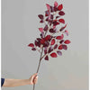 A hand holds a branch of autumn beech simulation flowers with deep red and purple leaves against a gray background.